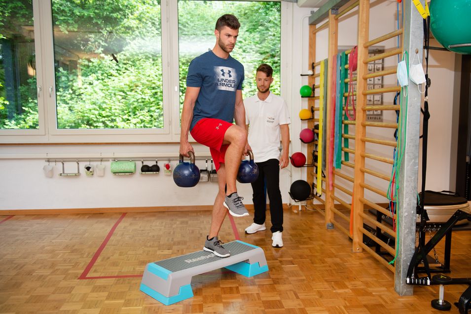 Patient mit Tape am linken Knie steht einbeinig und mit zwei Gewichten in den Händen auf einem Stepper und wird vom Physiotherapeuten beobachtet.