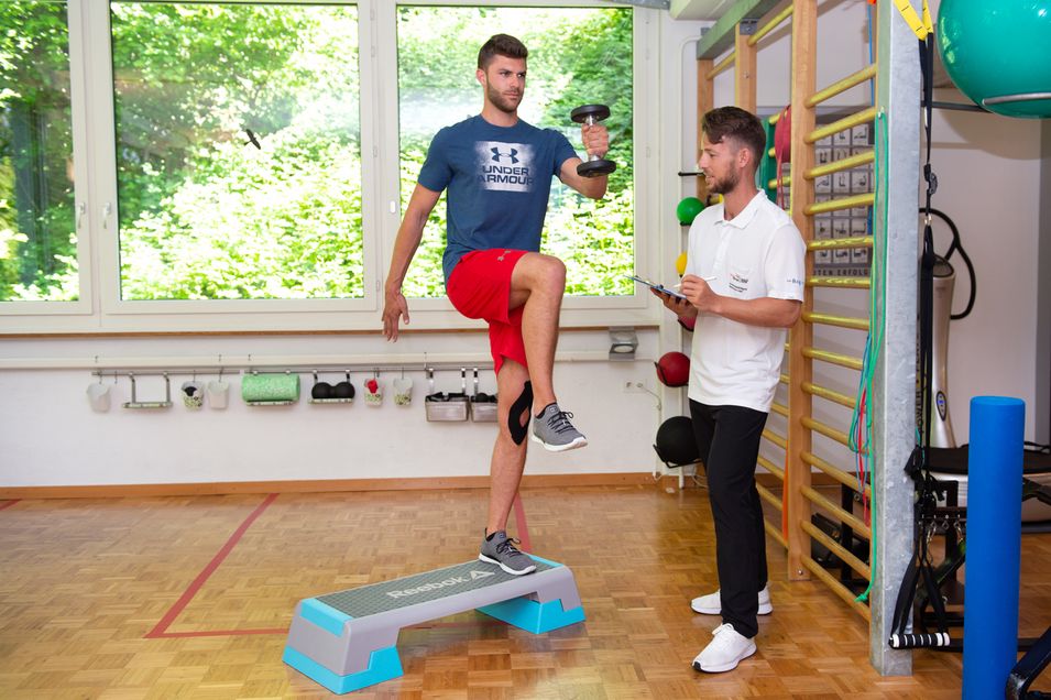 Patient with a tape on the left knee stands with one leg on a stepper with two weights in his hands. The right thigh points horizontally forward. The physiotherapist observes the execution.