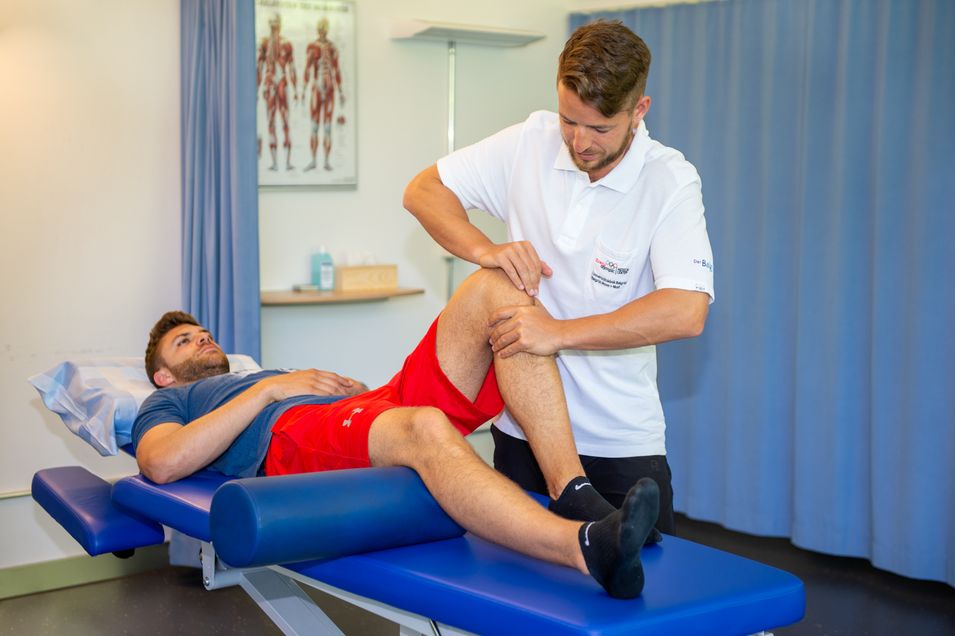 Patient is lying on his back on a massage bench with his left knee bent, while the physiotherapist is examining the patient's knee.