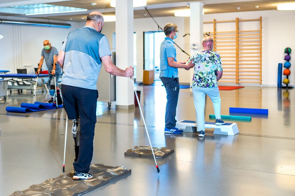 Three patients practice walking with prostheses and poles on uneven surfaces consisting of mats and steppers. They are supported by a physiotherapist.