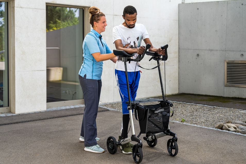 Patient mit einem laufunterstützenden Rollator wird beim Gehtraining von der Therapeutin von der linken Seite gestützt.