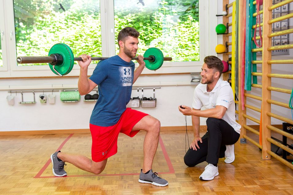 Patient with a barbell on the shoulder makes a lunge movement forward, the physiotherapist observes the execution.