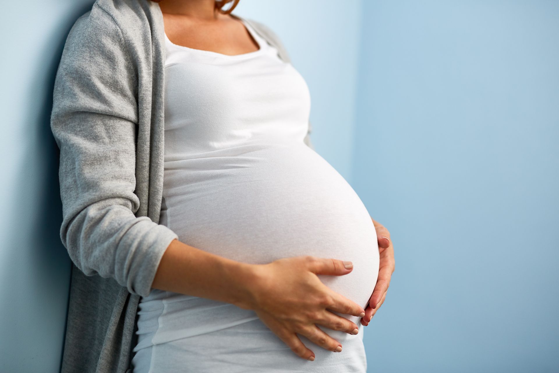 Oberkörper einer schwangeren Frau, die sich den Bauch hält und an der Wand anlehnt.