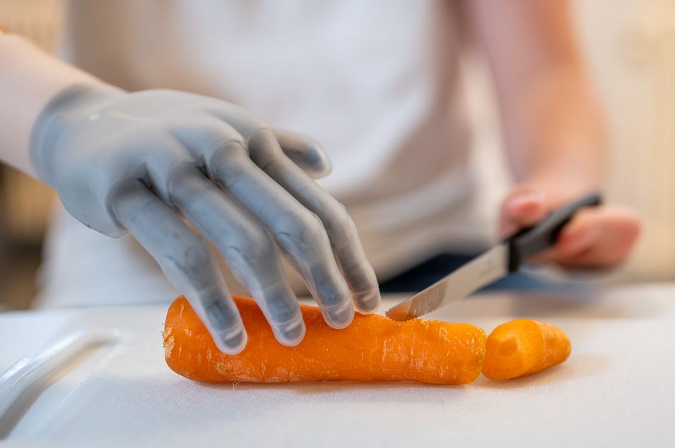 A man holds a carrot with his prosthetic hand while the other hand uses a knife.