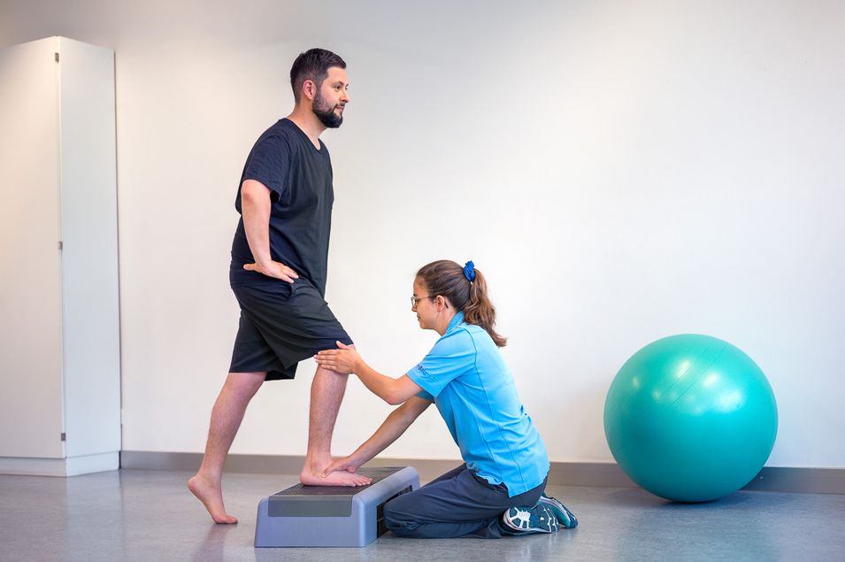 The patient stands with his right leg on a stepper and with his left leg on the floor. The physiotherapist examines the patient's left knee.