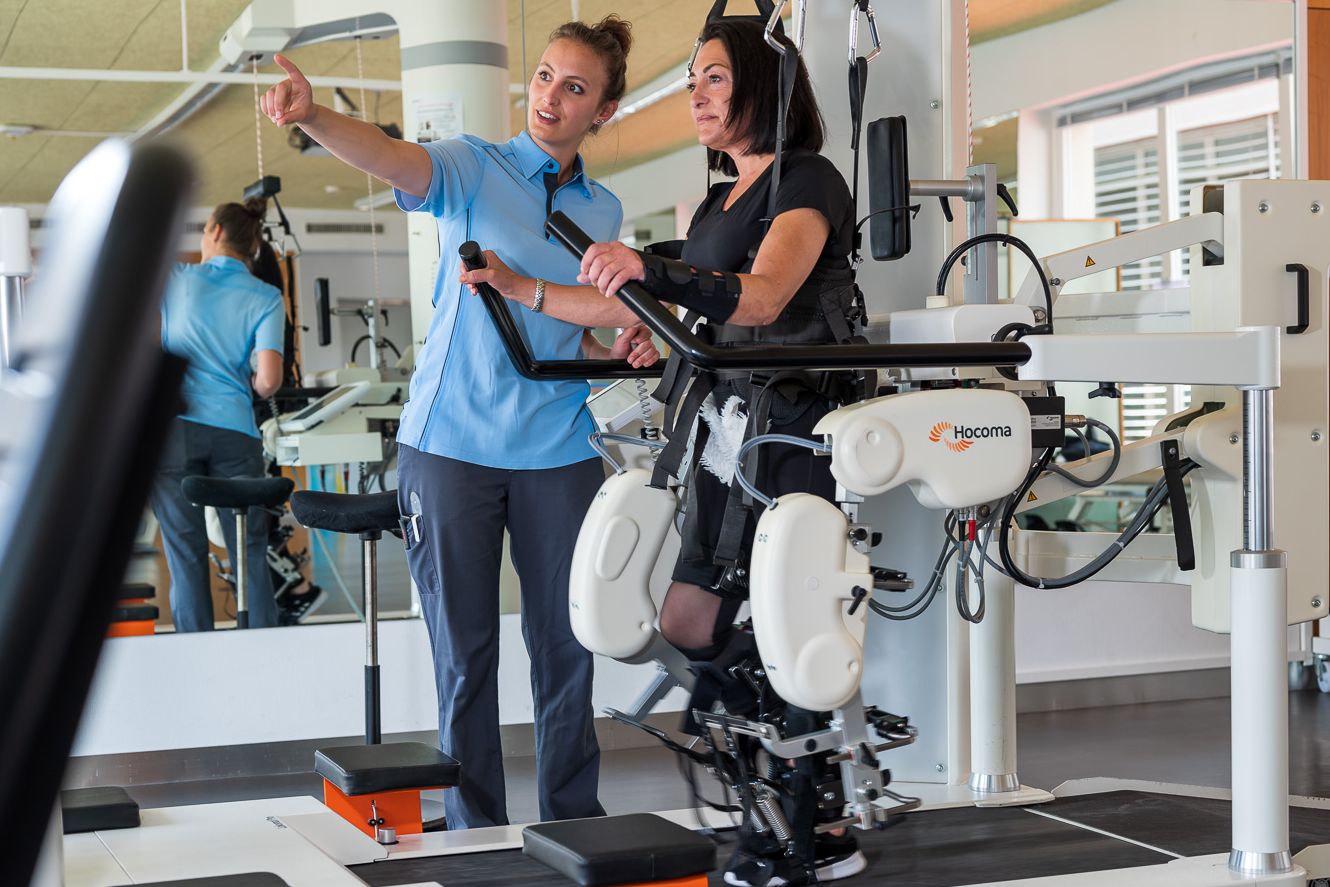 Patientin auf dem Laufband und bis zur Hüfte im Lokomat eingespannt, wird von der Physiotherapeutin instruiert.