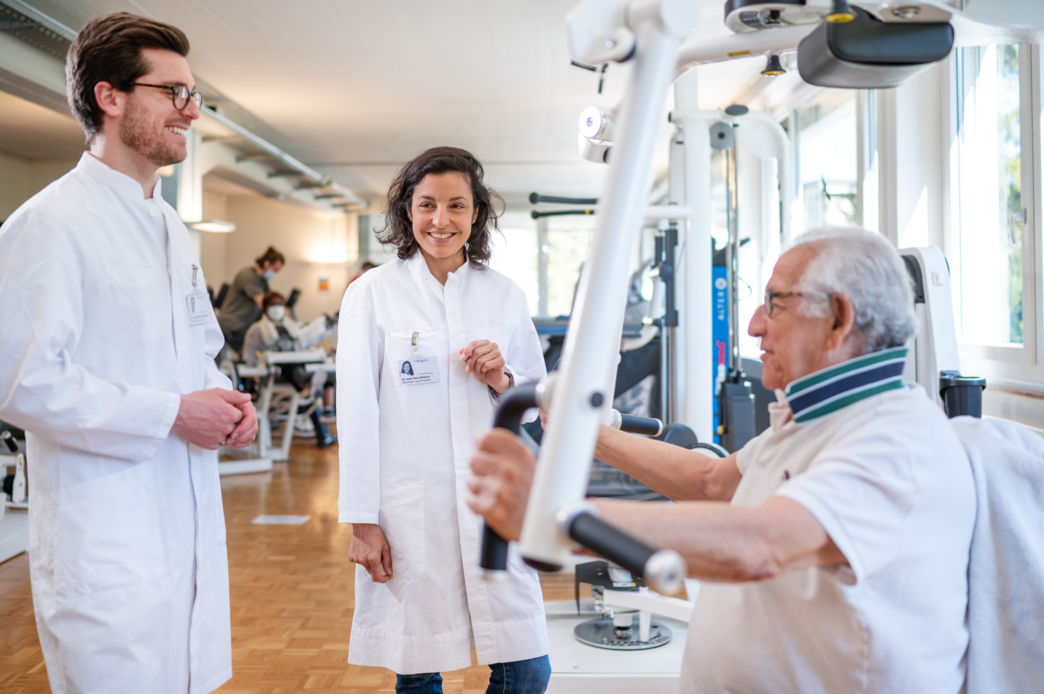 A male and female sports physician consult with a patient exercising on a piece of fitness equipment.