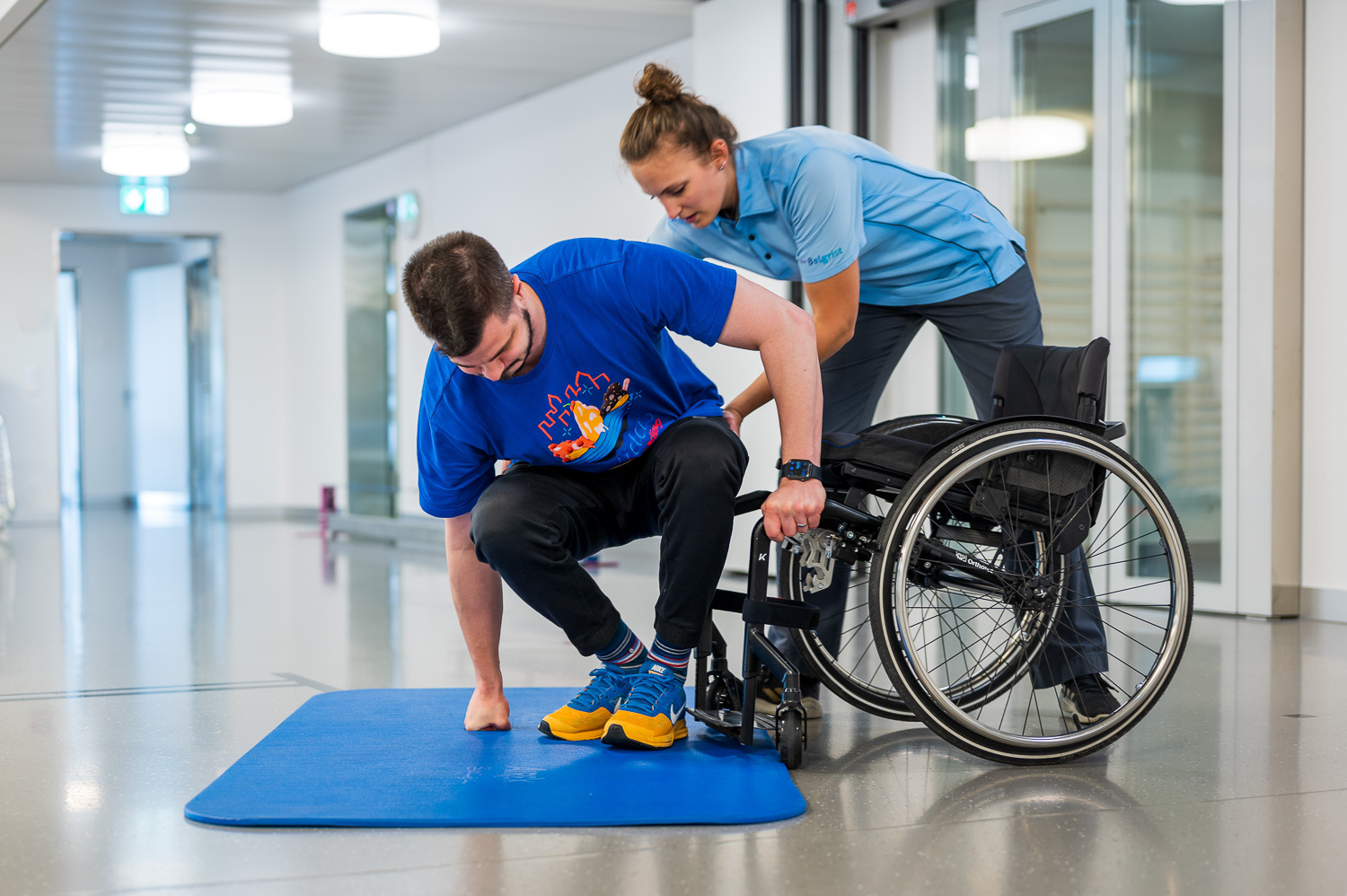 Paraplegic patient tries to get into the wheelchair backwards from a blue mat lying on the floor. The physiotherapist helps him and holds him by the pelvis.