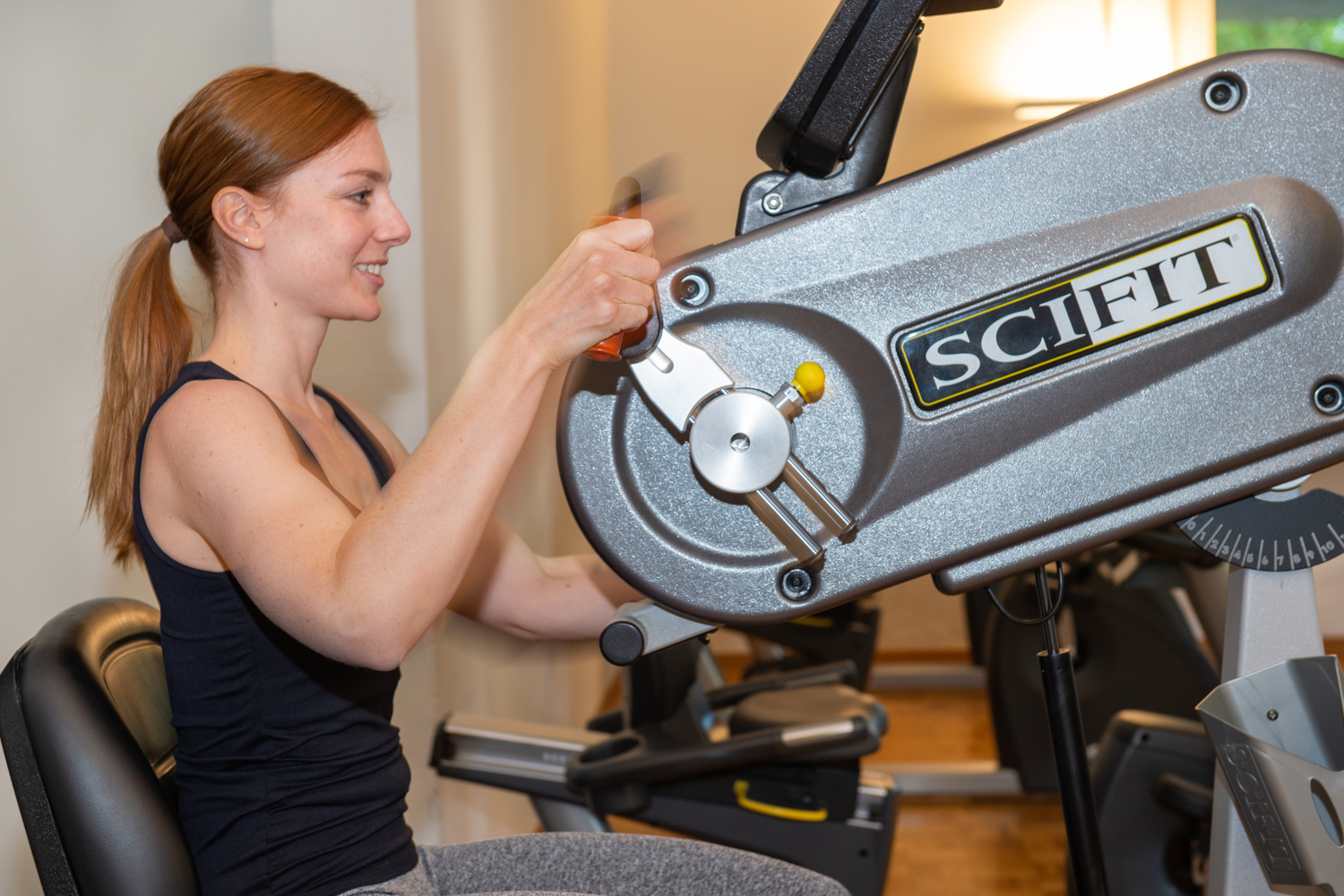 Patient with long brown hair and black top sits at a walking ergometer and makes circular movements while smiling.
