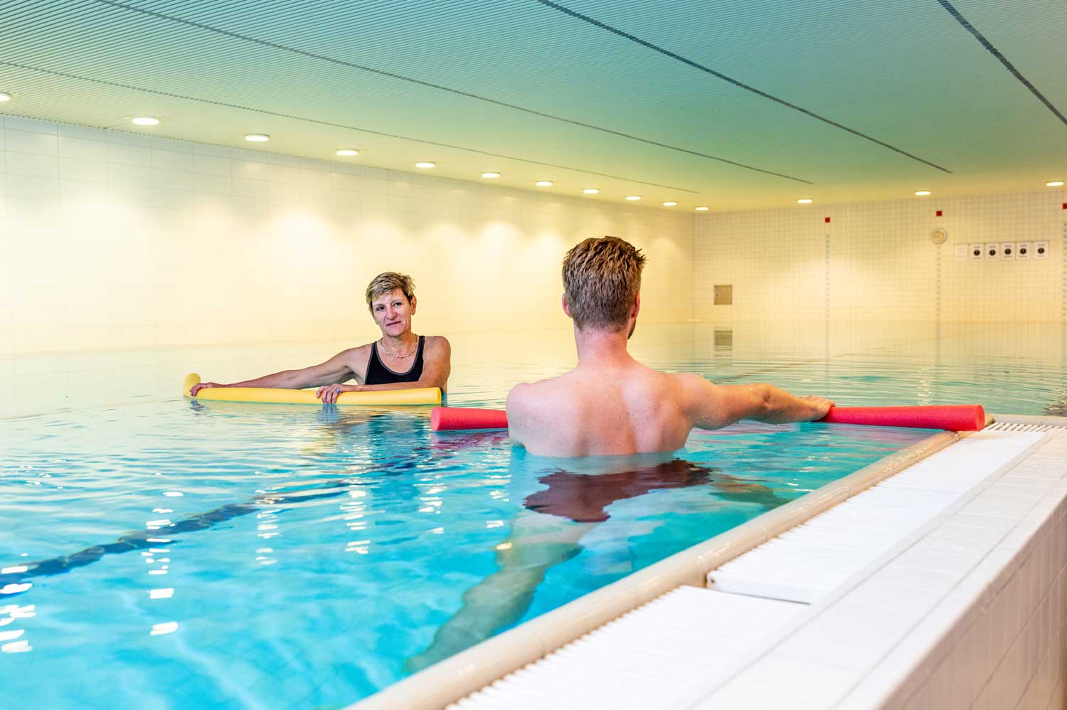 Eine Patientin und ein Patient befinden sich bis unterhalb der Schulter im Wasserbecken und machen eine Oberkörperdrehung. Gleichzeitig haben sie eine Wassernudel in den Händen, die an der Wasseroberfläche entlang bewegt wird.