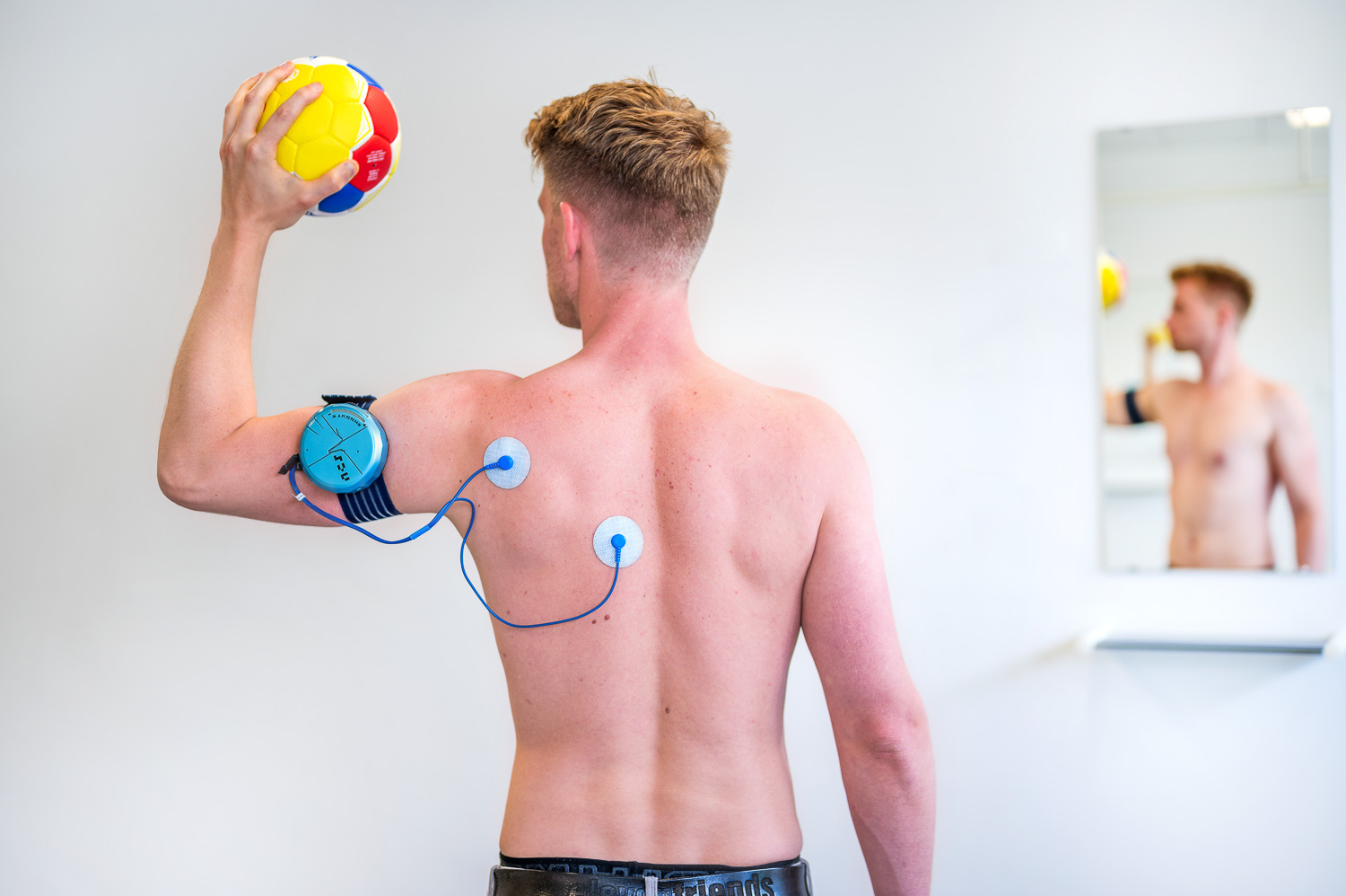 Patient is seen from behind with the Shoulder Pacemaker on his left shoulder, holding a handball.