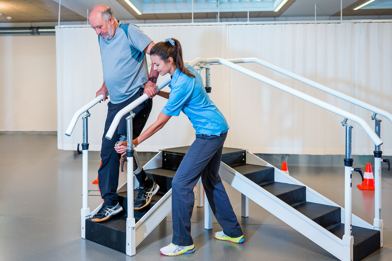 Patient mit einer Oberschenkelamputation am linken Bein läuft mit Hilfe der Physiotherapeutin eine zu Übungszwecken aufgestellte Treppe runter.