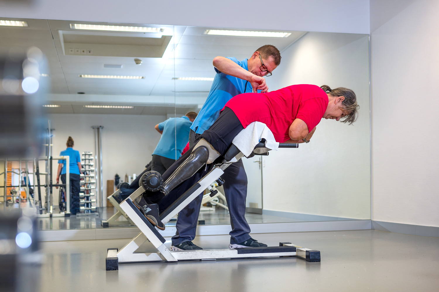 Patientin mit einer Unterschenkelprothese ist in einer schräg nach oben gerichteten Position und macht mit Unterstützung des Physiotherapeuten Rückenübungen, indem sie den Oberkörper entgegen der Schwerkraft nach oben hebt, während ihr Unterkörper bis zur Hüfte fixiert ist.