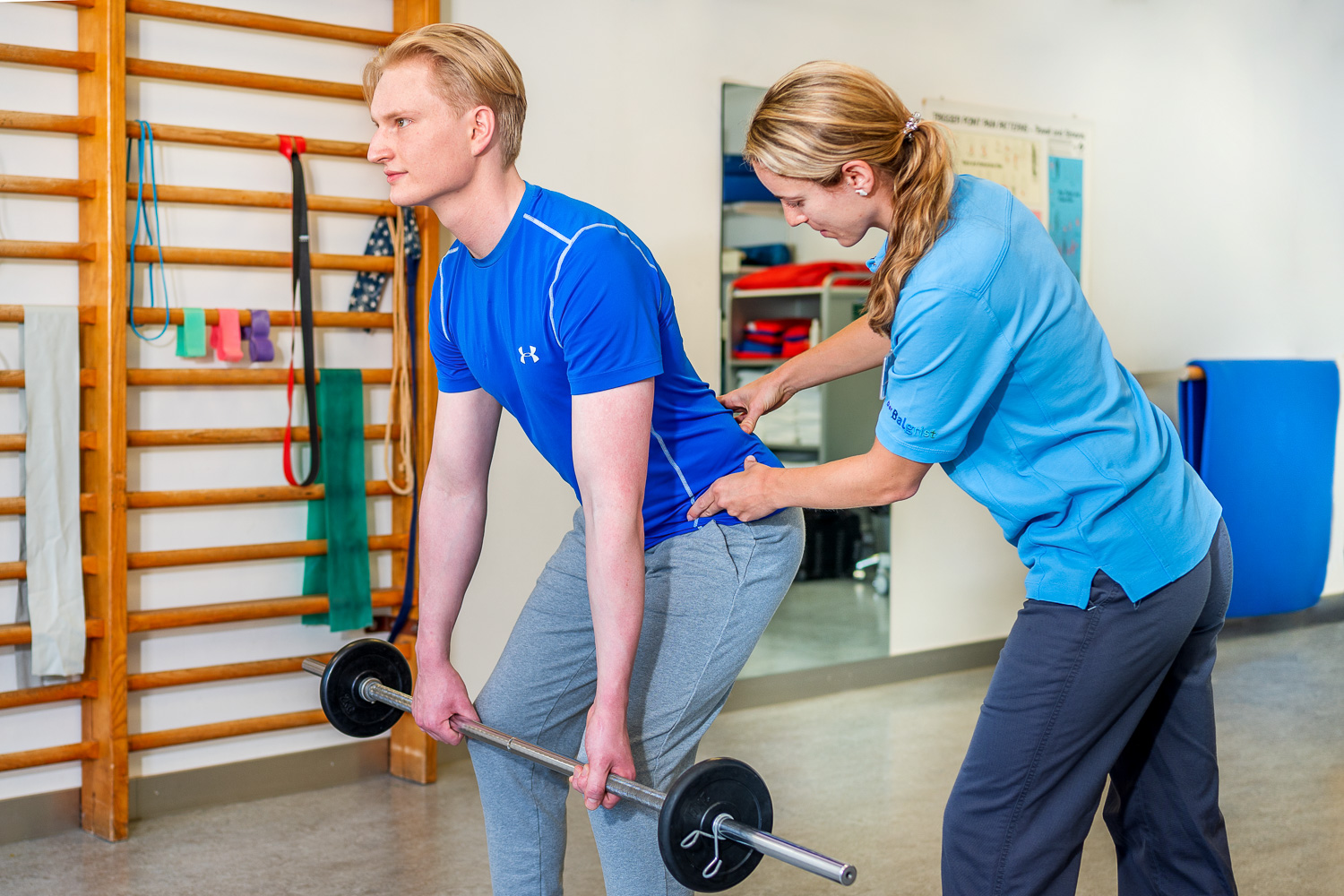 The physiotherapist pays attention to the correct posture of the patient, who performs the back exercise deadlift with a barbell.