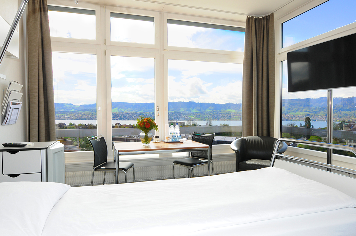 Bed in the foreground of a private room with a view of Lake Zurich.